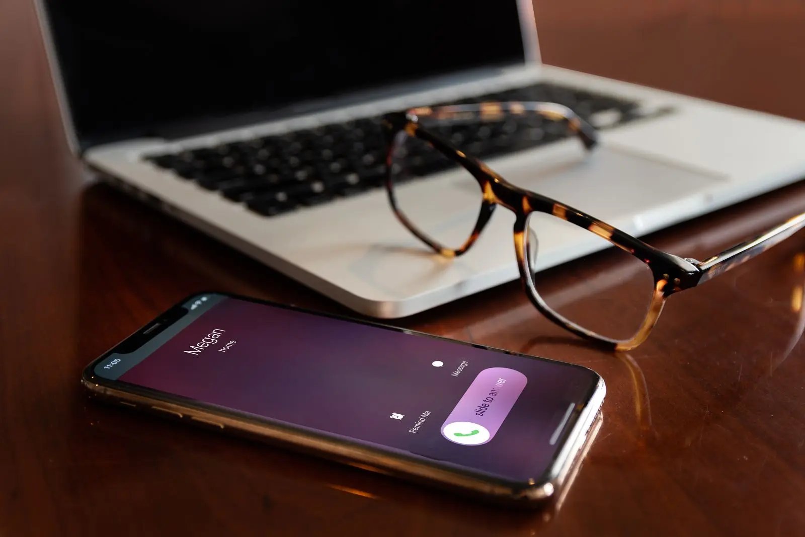 Smartphone on a desk showing an incoming call, next to a laptop and glasses